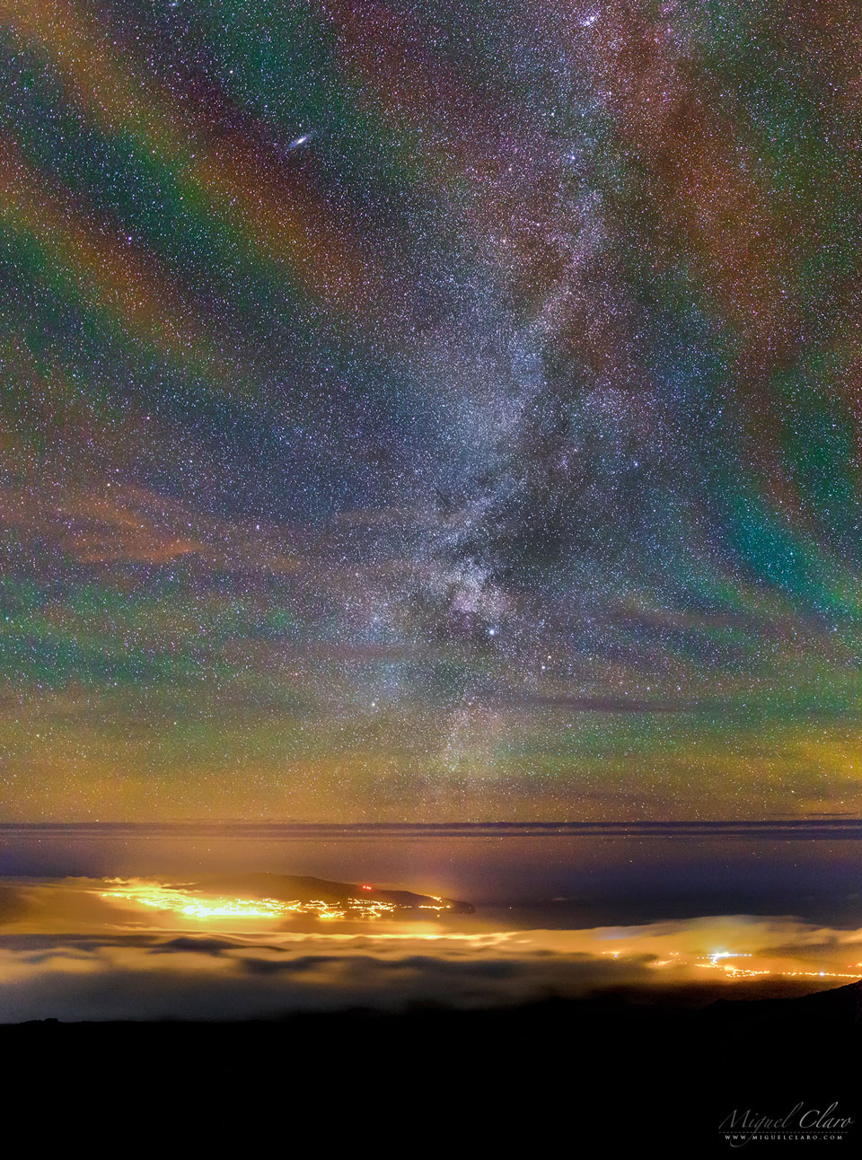 A starfield is seen over water, clouds, and the lights of a city below. The starfield is oddly not black, but shows a repeating assortment of transparent colors diagonally from the lower left to the upper right. Please see the explanation for more detailed information.