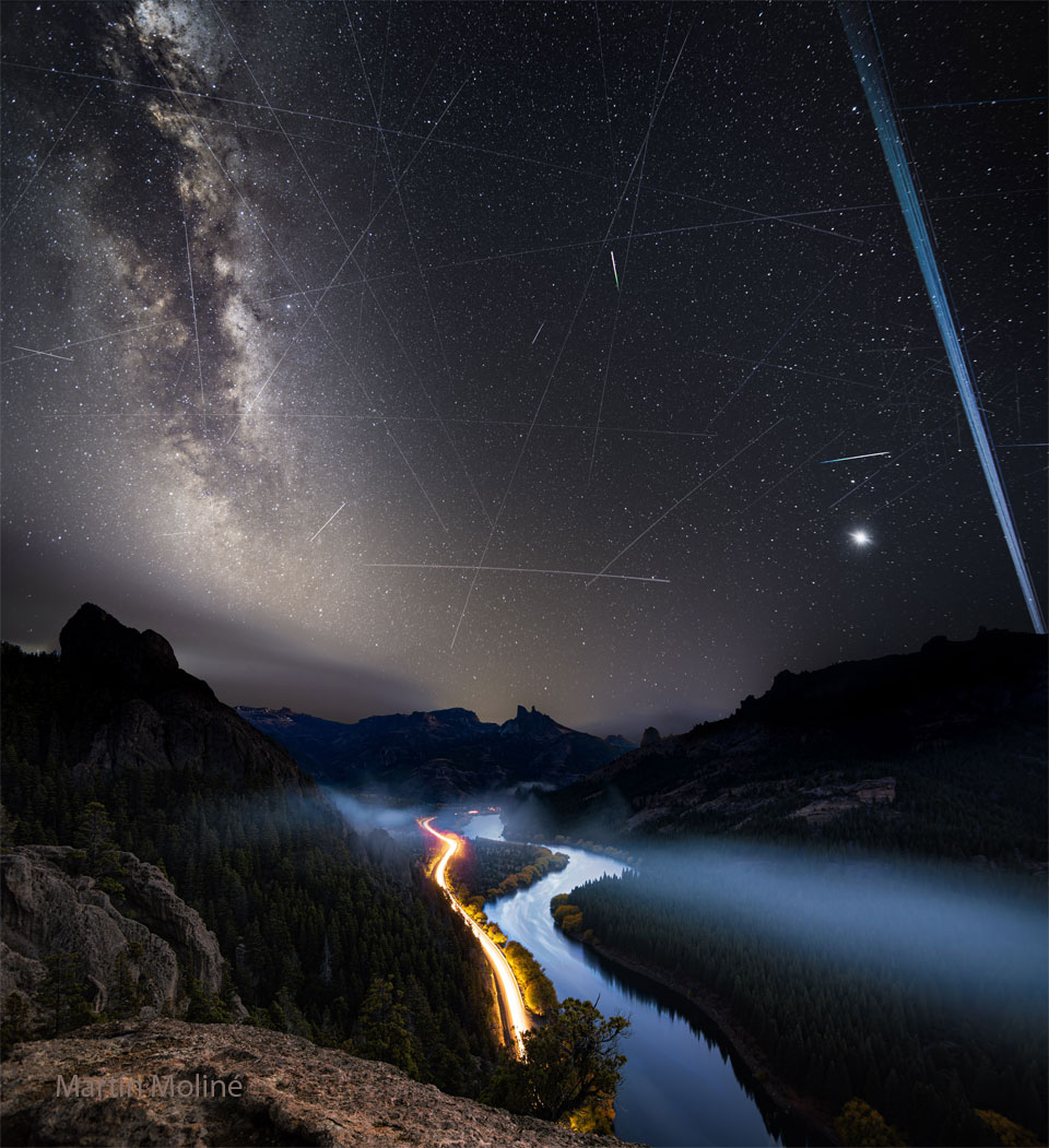 A night sky is shown with many stars and streaks. In the foreground at the bottom are hills, a river, and the red and white streaks of car lights. Please see the explanation for more detailed information.