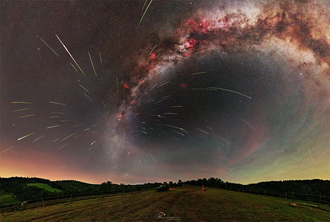 A starfield is shown above a grassy field with hills on the horizon. The band of our Milky Way Galaxy arches across toward the right. Many streaks appear emanating out from a place on the Milky Way just above the horizon. Please see the explanation for more detailed information.